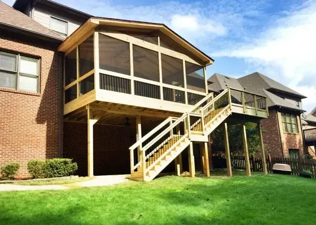 A wooden screened-in porch with an attached deck and staircase overlooking a green backyard at a brick house.