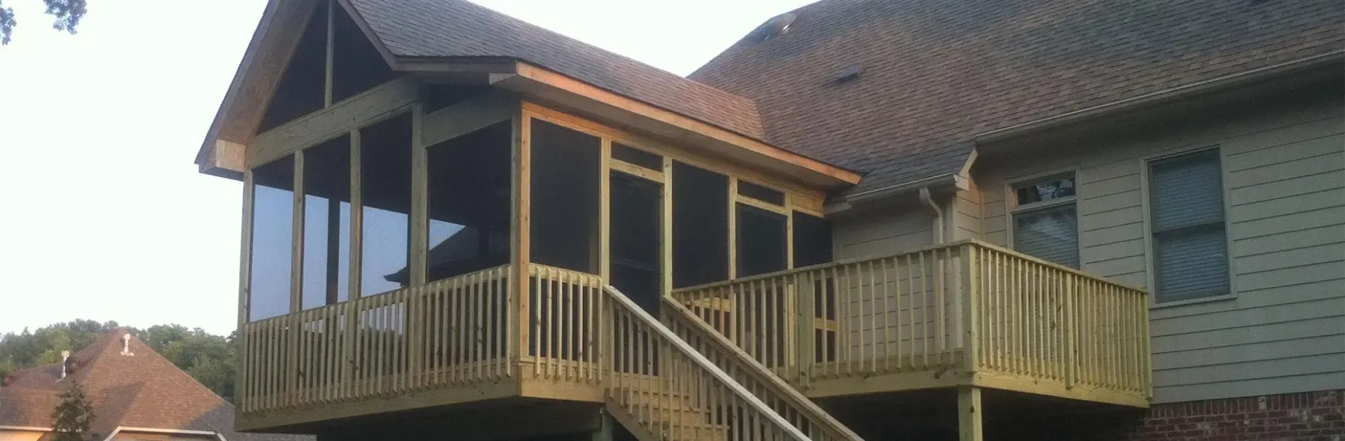 A raised wooden deck featuring a screened-in porch with a gable roof attached to the rear of a tan-sided house.