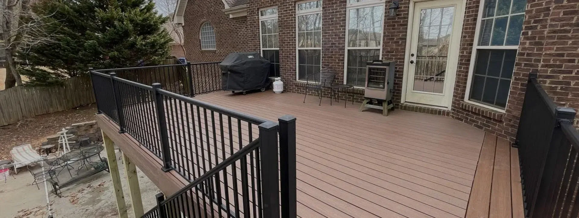 A brown composite deck with a black railing, featuring a covered grill and outdoor seating against a brick house wall.