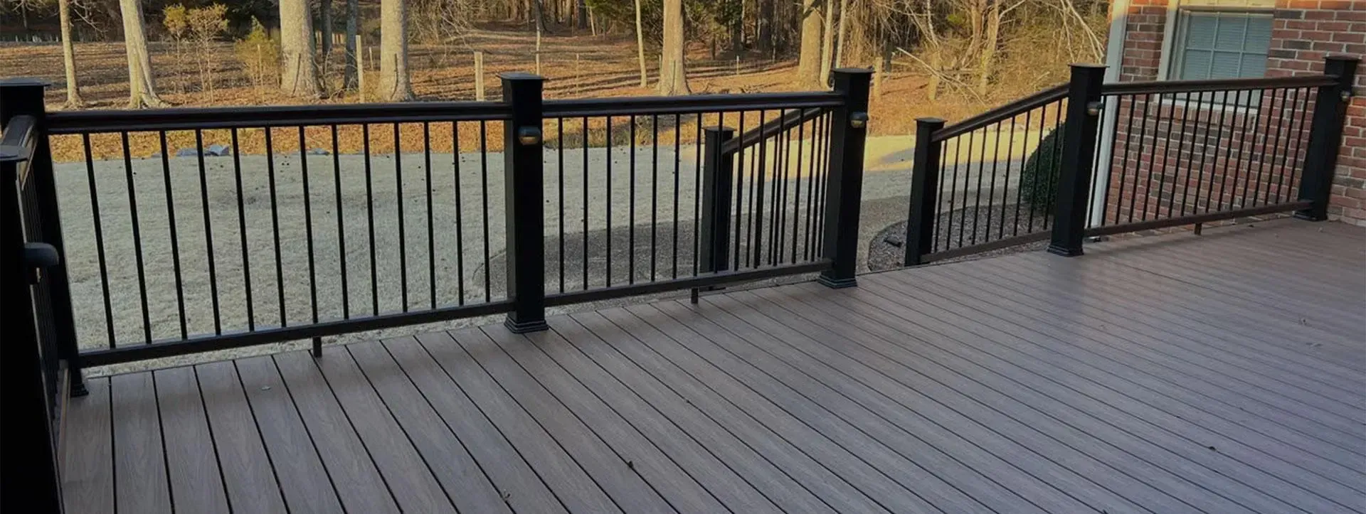 A wooden deck with a black metal railing overlooking a wooded area at sunset.