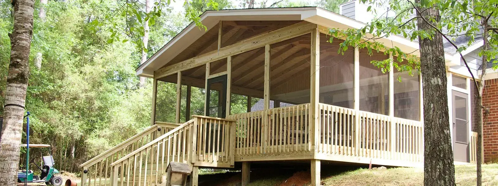A light wood screened-in porch with a peaked roof and stairs, set in a wooded area with mature trees.