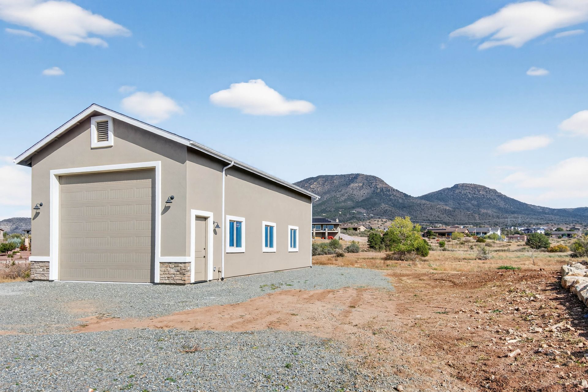 A beige utility shed is on a barren area, close to a residence.