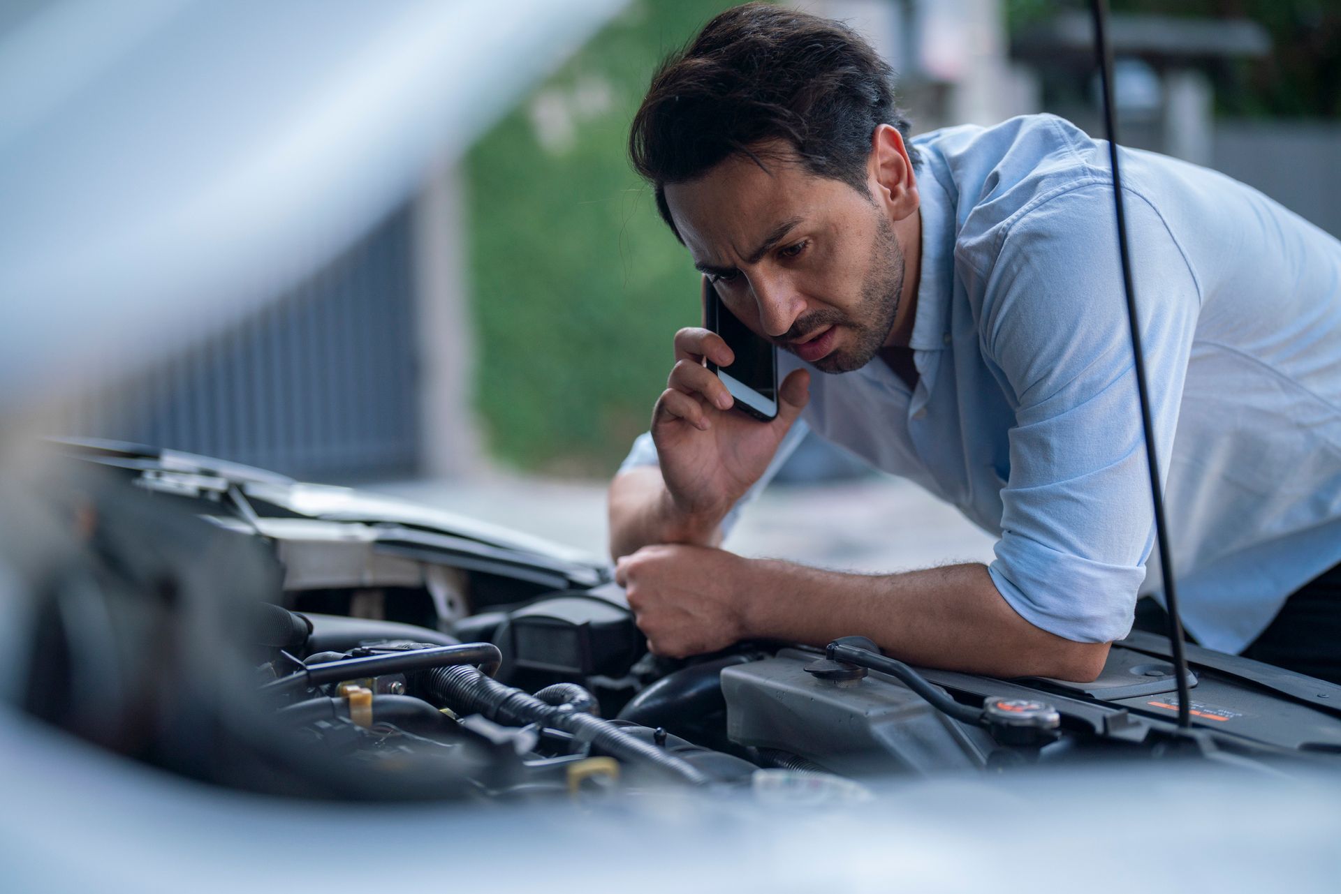 Image of a man checking under a car hood while calling on his smartphone.