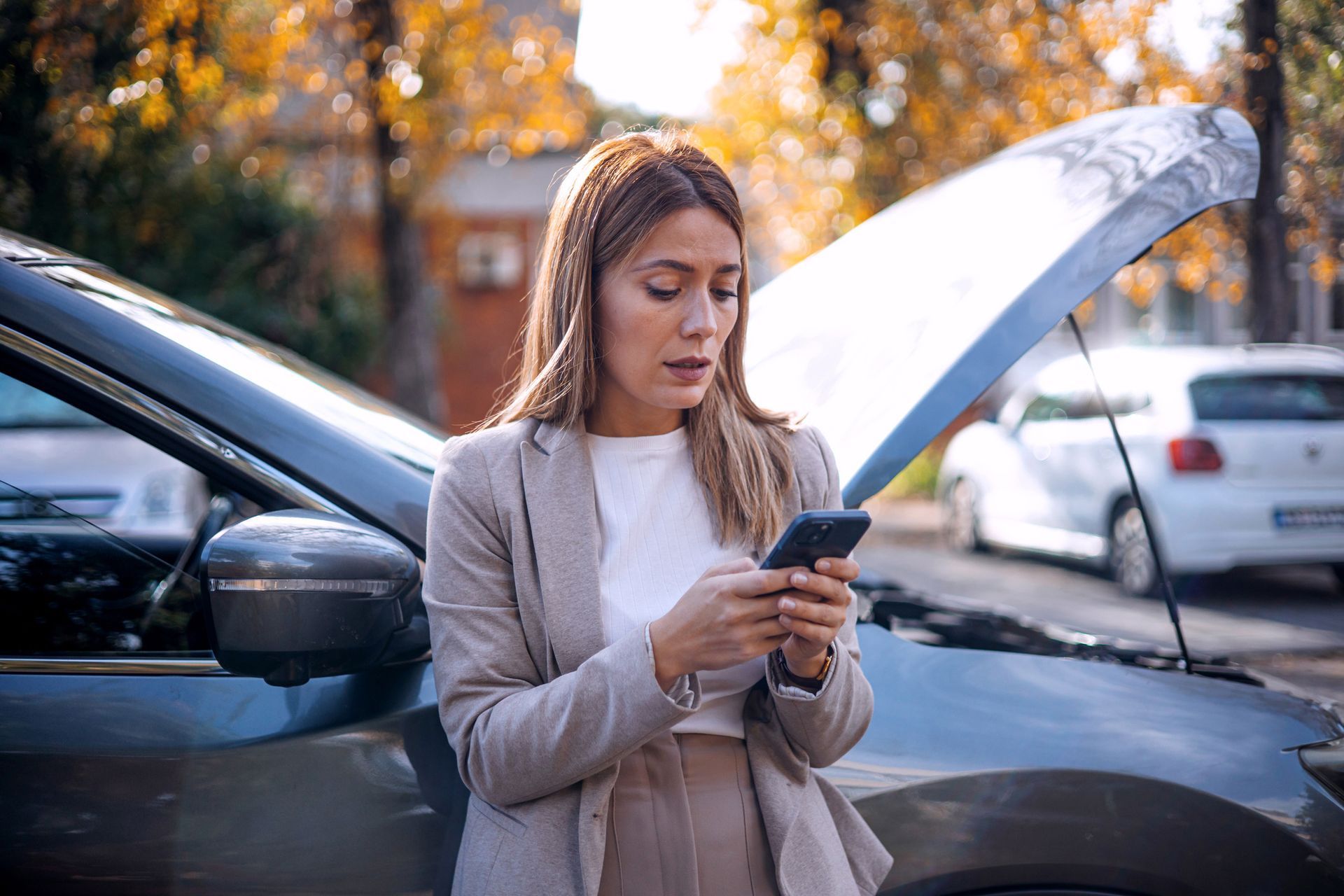 A woman on her phone beside car with open hood on roadside.