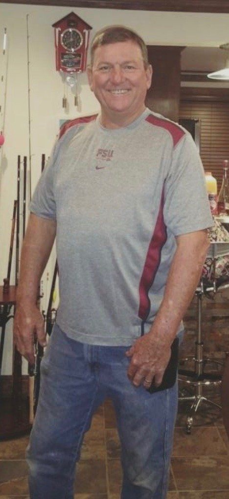 Man wearing a gray shirt and jeans smiles indoors, near a clock and pool cues.