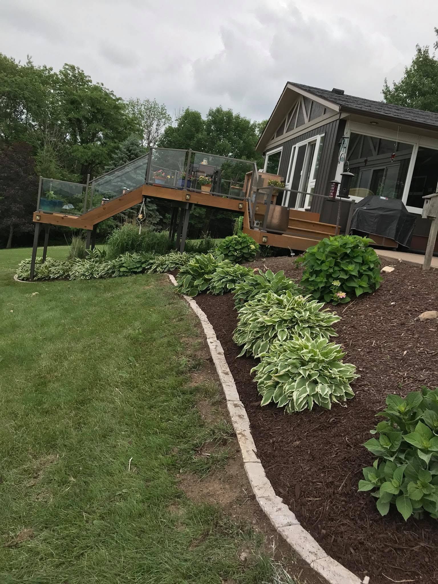 Landscaped yard with mulch beds, curved stone edging, and a house deck with stairs in the background