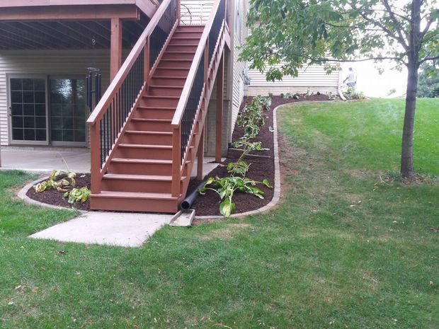 Wooden staircase to a deck beside a house, with landscaped beds and a grassy yard.