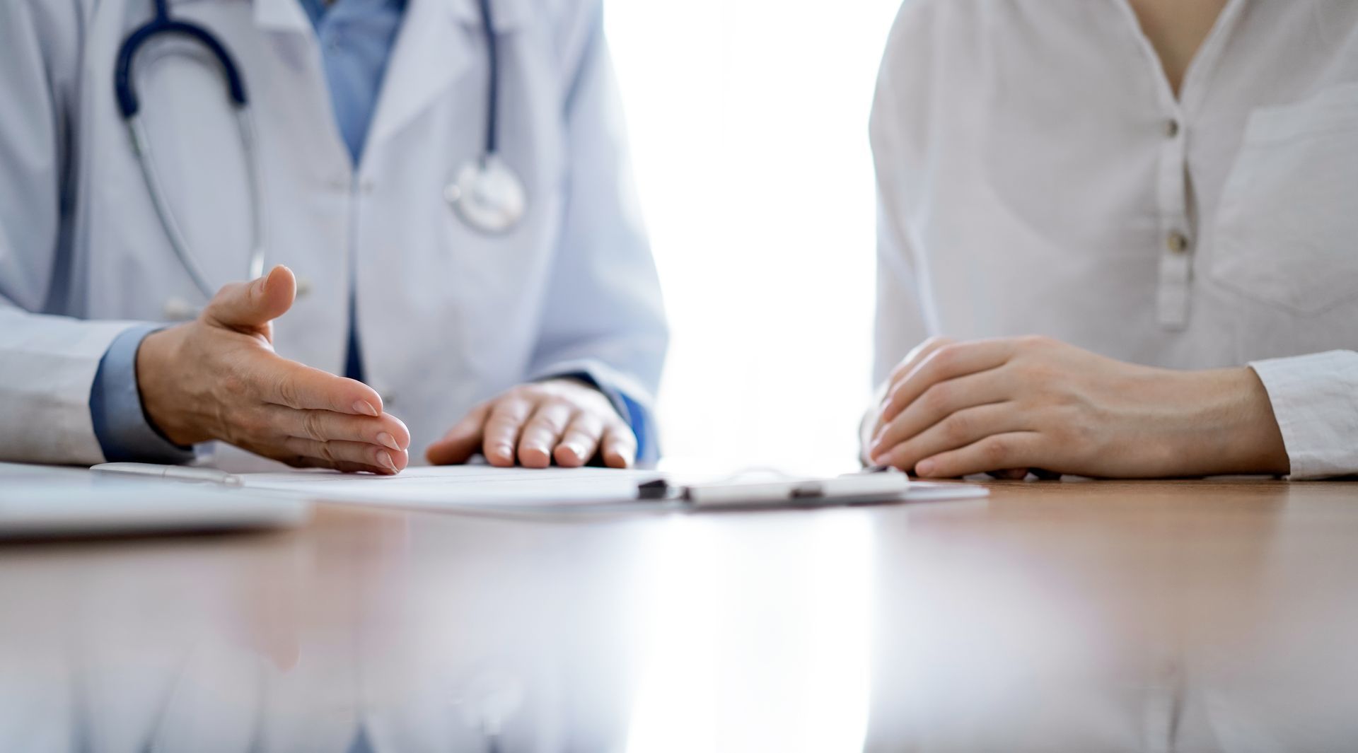 Un medico in camice bianco gesticola mentre discute di documenti con un paziente in camicia bianca seduto a un tavolo.
