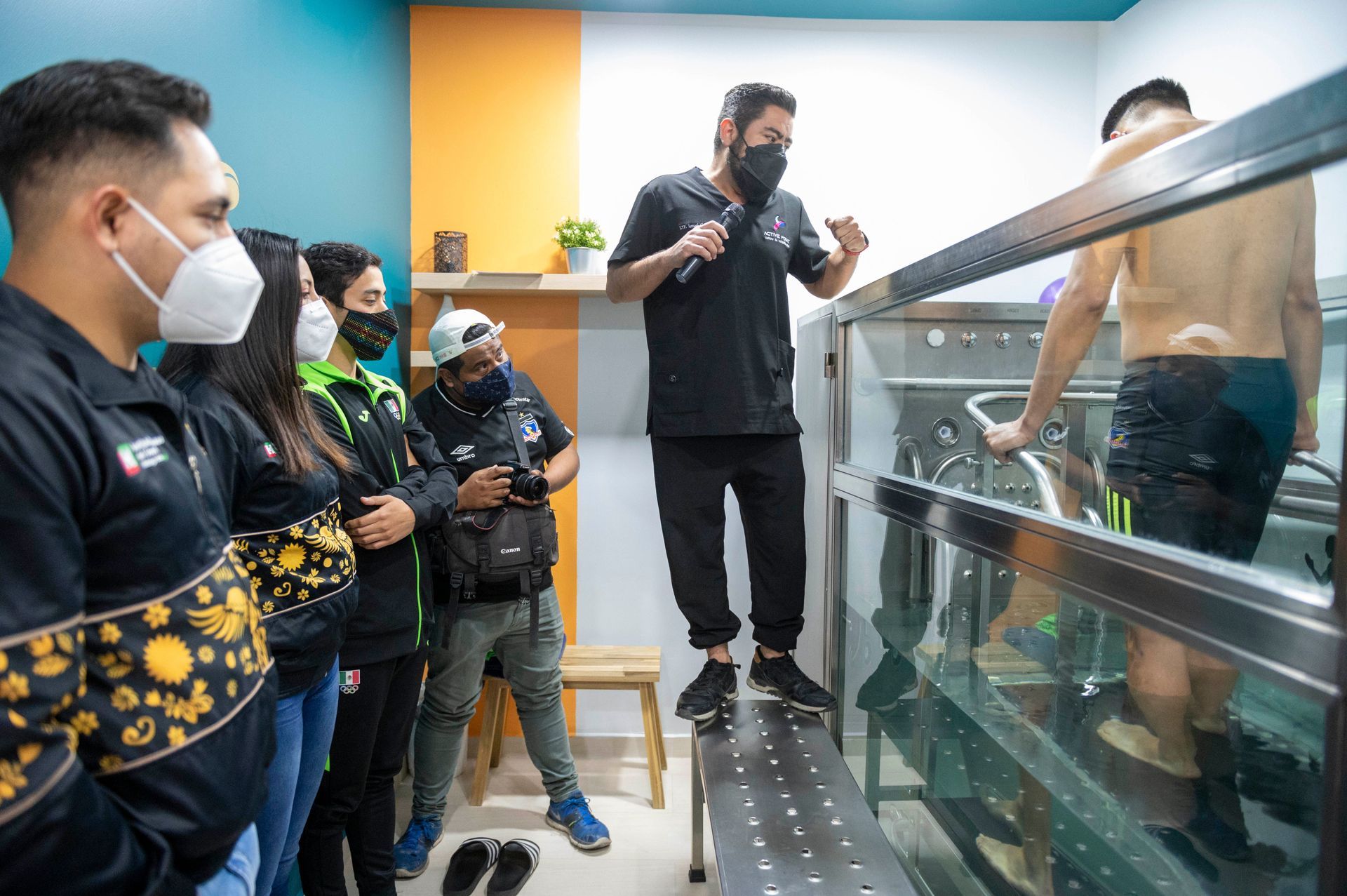 Personas observando a un hombre en una cinta de correr acuática mientras el instructor habla. La sala tiene paredes coloridas.