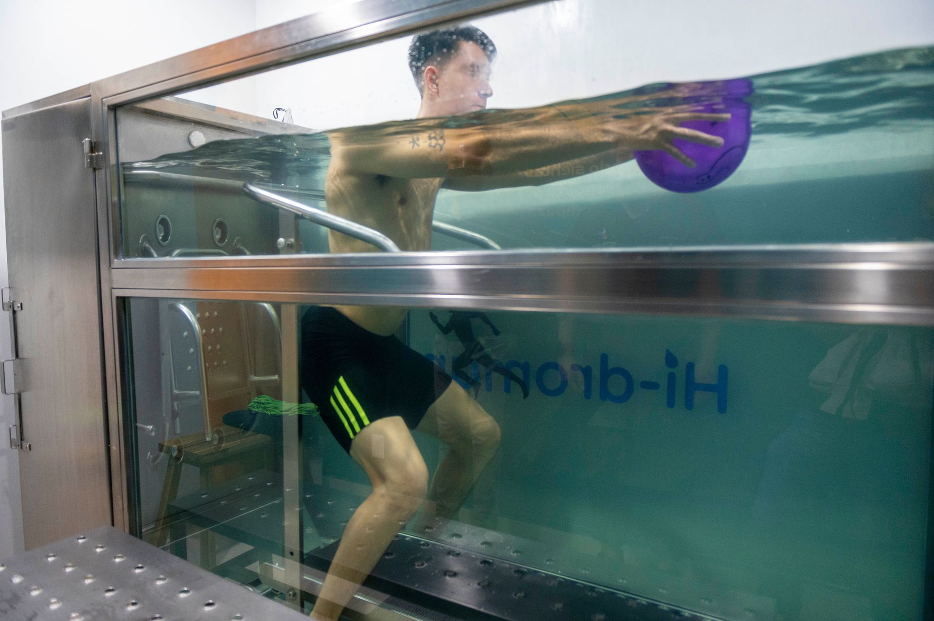 Hombre haciendo ejercicio bajo el agua en una piscina terapéutica, sosteniendo un disco morado. Lleva pantalones cortos negros.