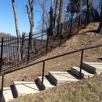 Concrete steps with a handrail ascend a grassy hill towards a black metal fence and trees, with water in the background.