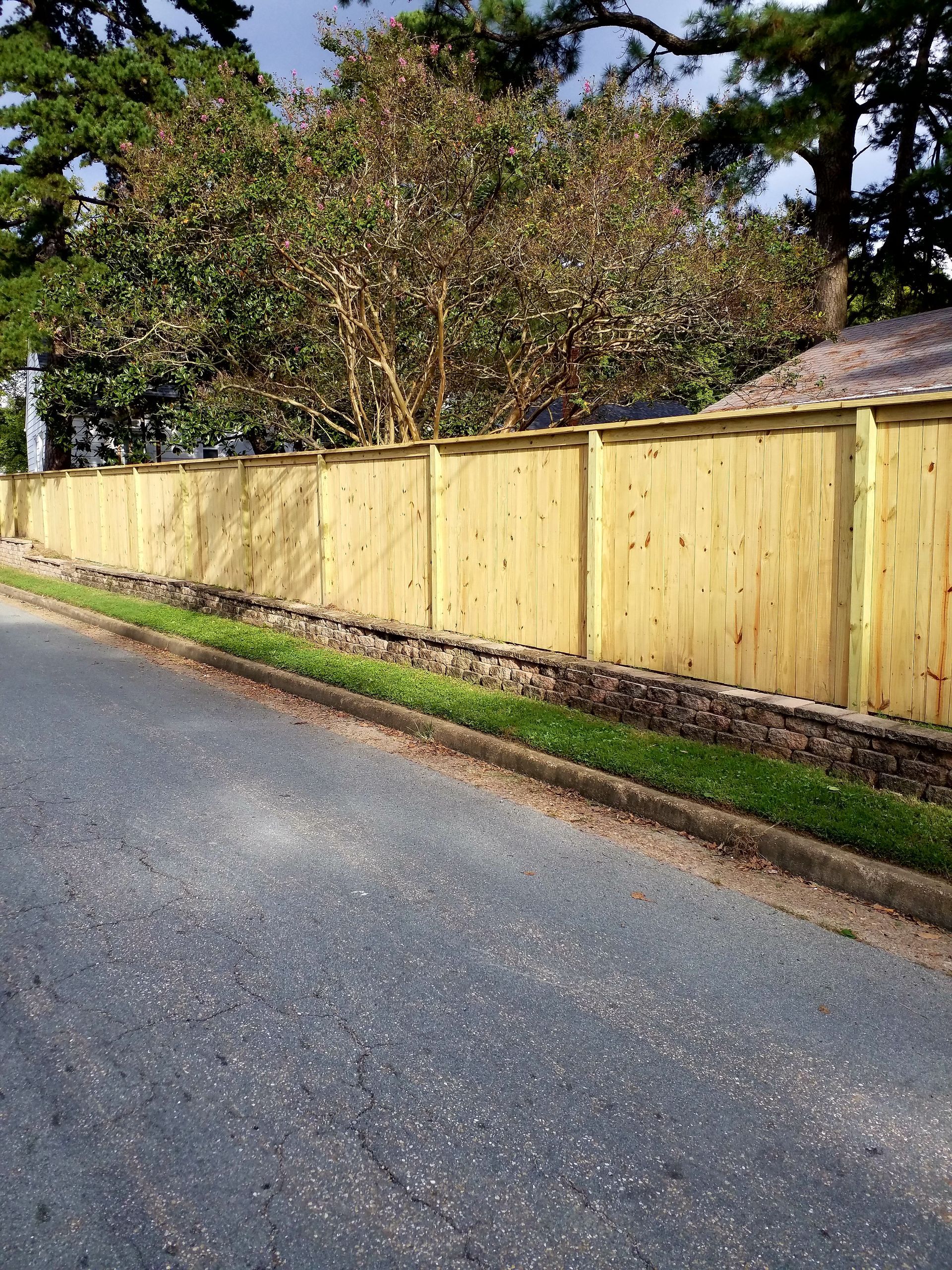 New wooden fence along a street; light-colored wood, green grass, and trees in the background.