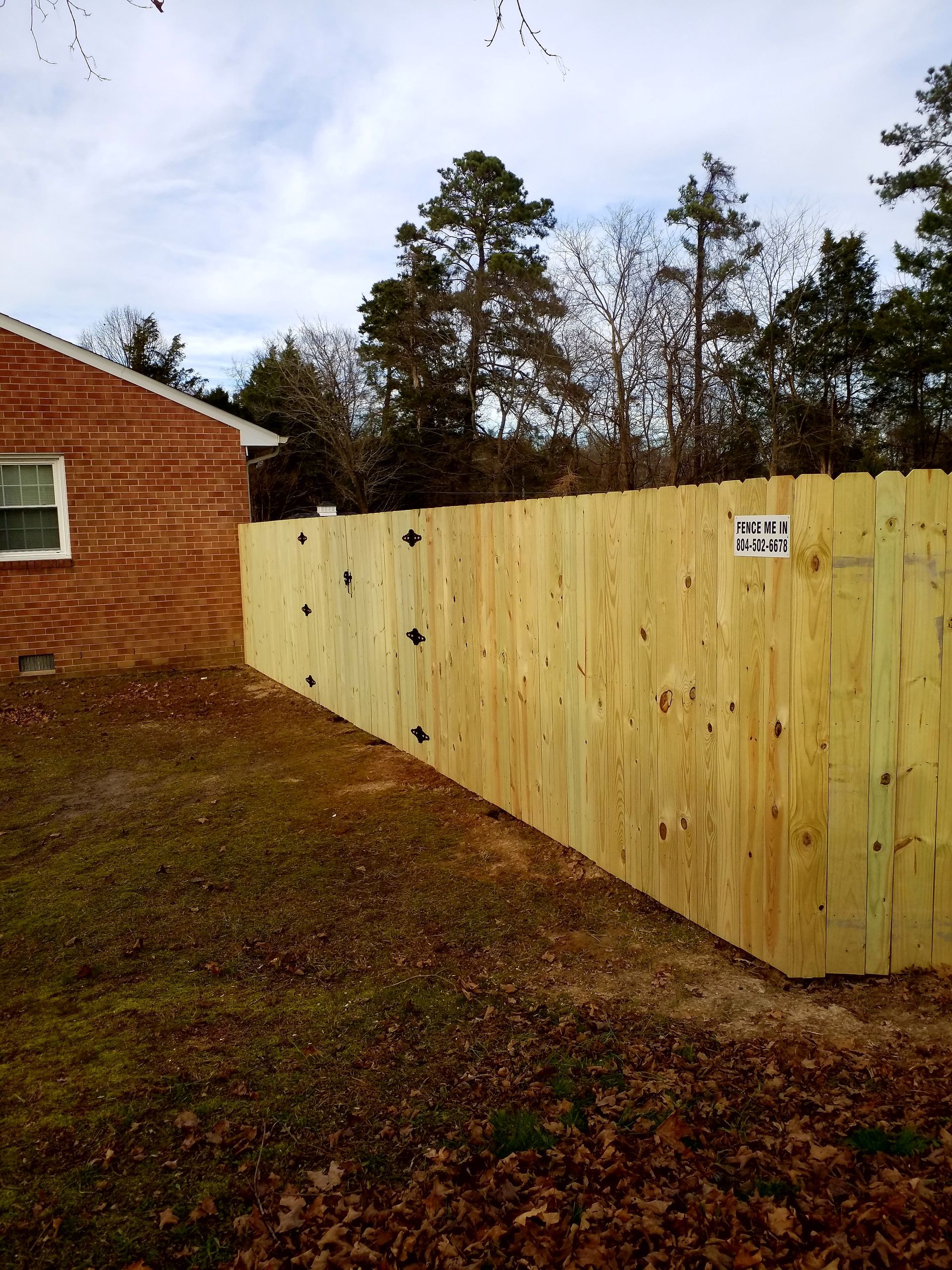 A new wooden fence next to a brick building and a grassy yard with trees in the background.
