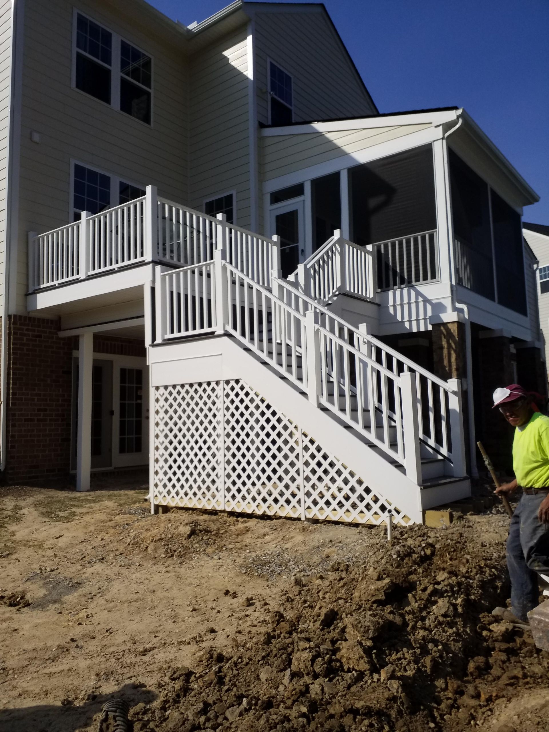 White deck and screened porch attached to a two-story beige house. Person stands nearby, on dirt.
