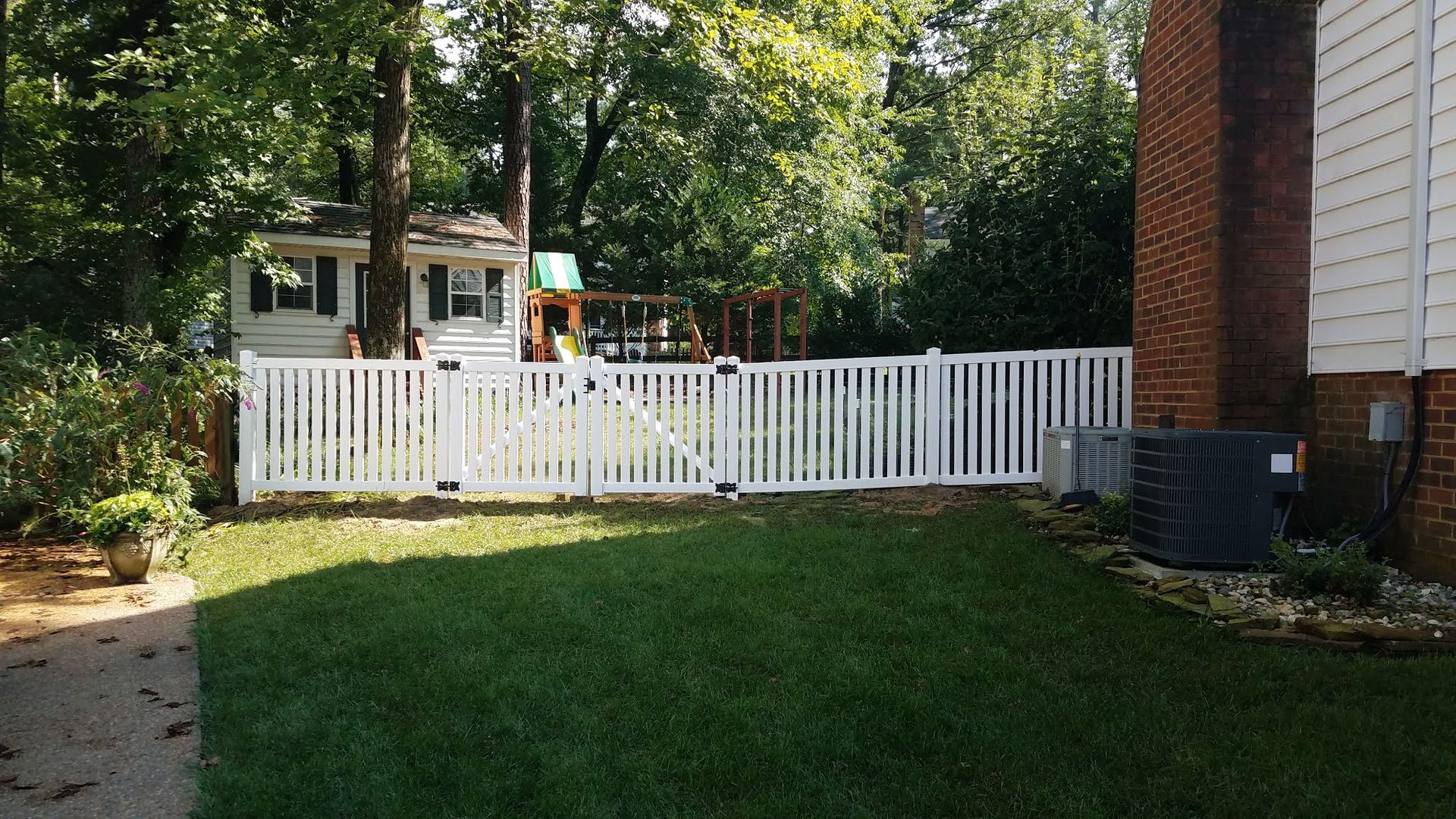 White picket fence encloses a backyard with a playset and house, a brick wall is to the right.