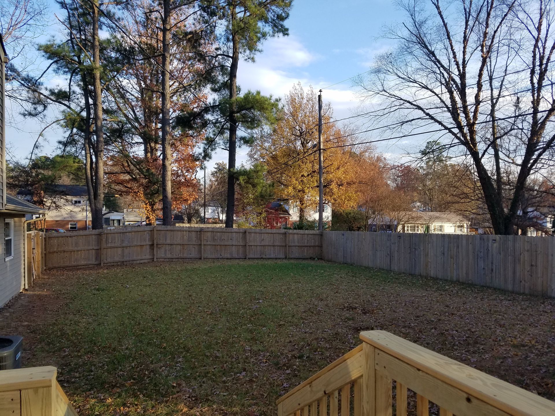 Wooden fenced backyard with green grass, trees, and a wooden deck. Autumn leaves.