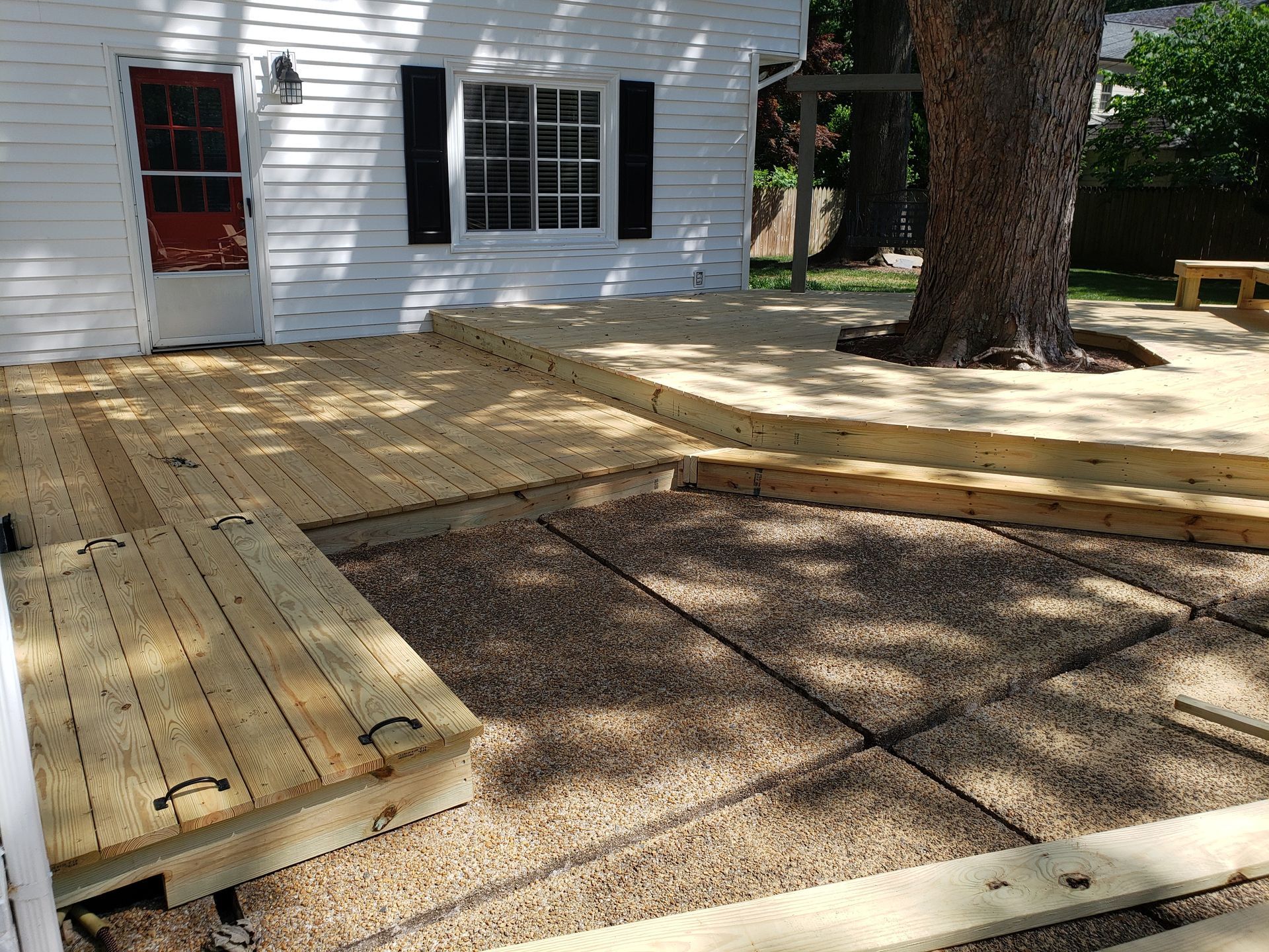 Wooden deck with pebble ground, beside a white building with a tree.