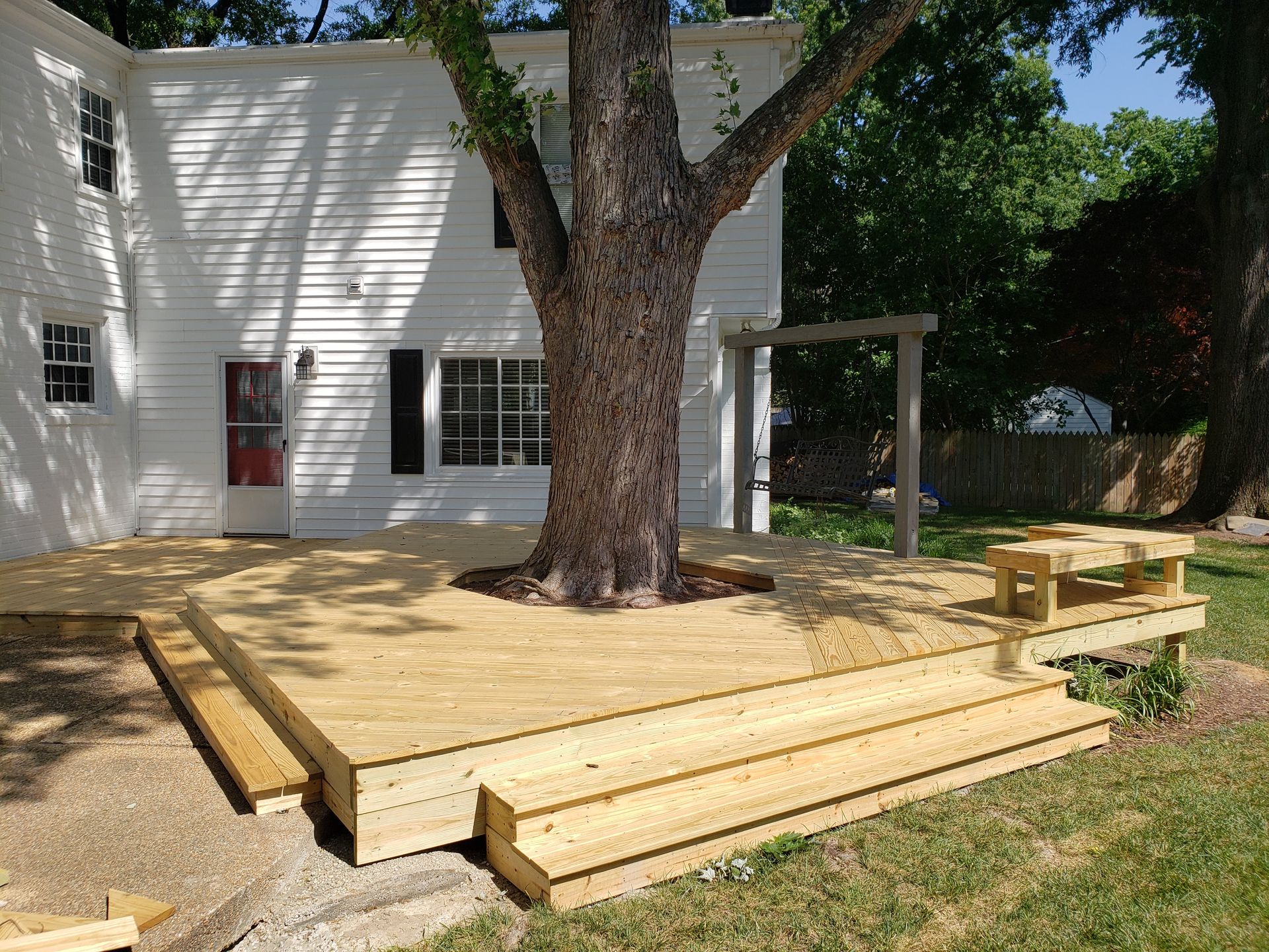Wooden deck wraps around a large tree, with a small bench nearby, and a white house in the background.