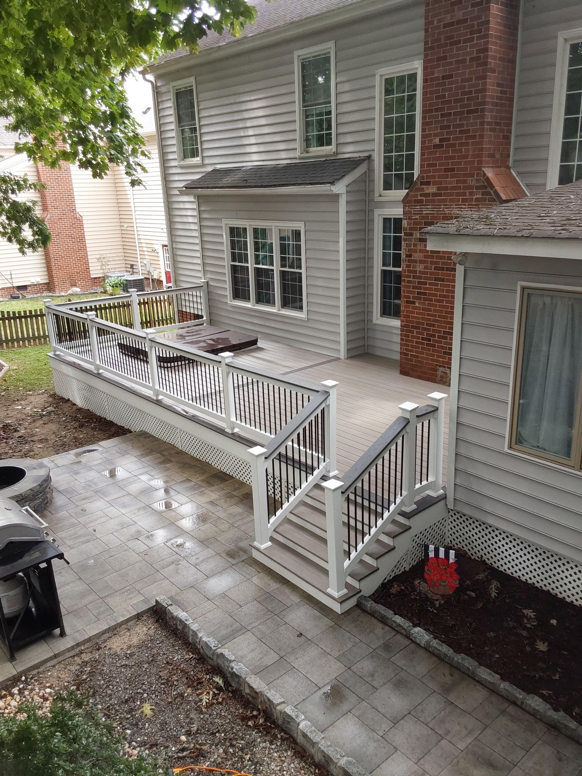 Backyard patio with stairs, deck, and gray siding home. White railings, stone path, and brick chimney visible.