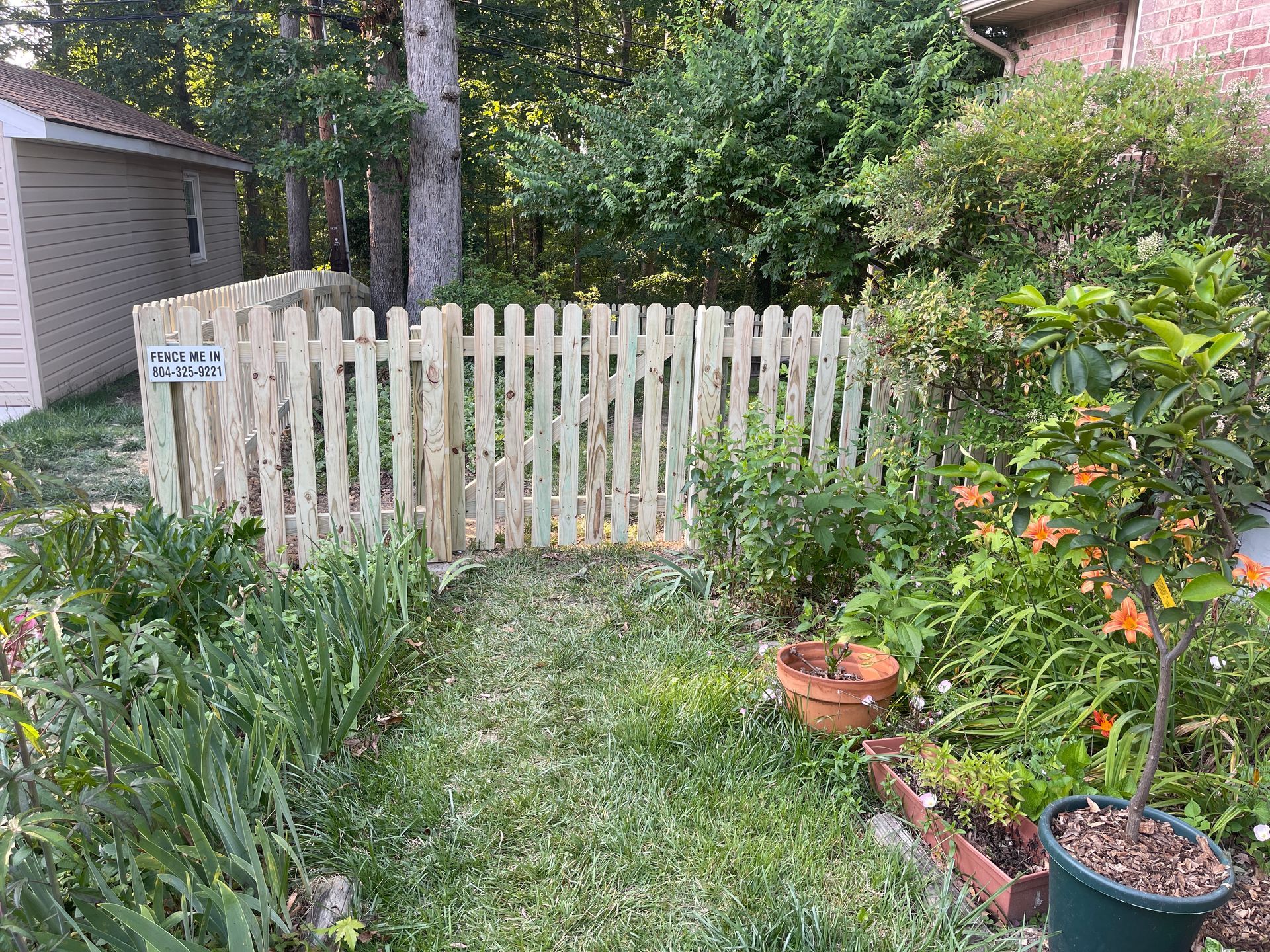 A wooden picket fence encloses a garden area with plants and flowers, near a shed and trees.