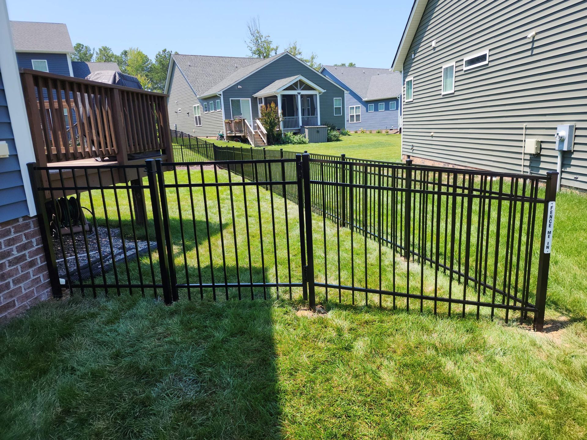 Black metal fence encloses a green lawn, bordering a deck and a house. Sunny day.