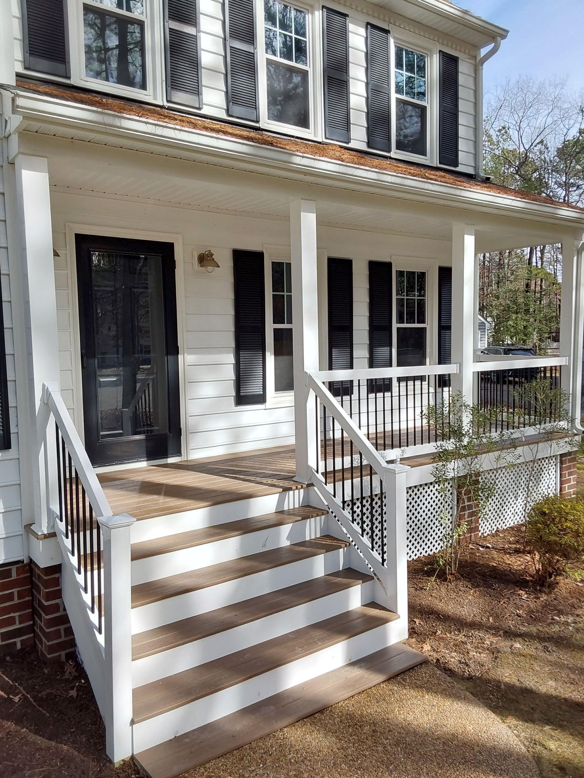 White house with porch, black door and shutters, brown steps, and landscaping.