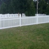 White picket fence bordering a grassy lawn, with trees in the background.