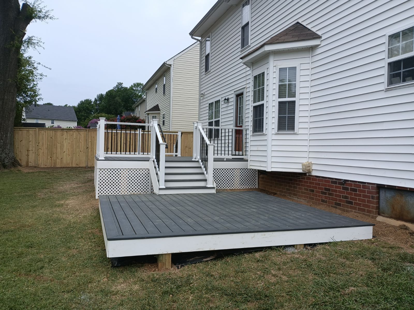 Gray deck with stairs and railing attached to a white house with a bay window; backyard setting.