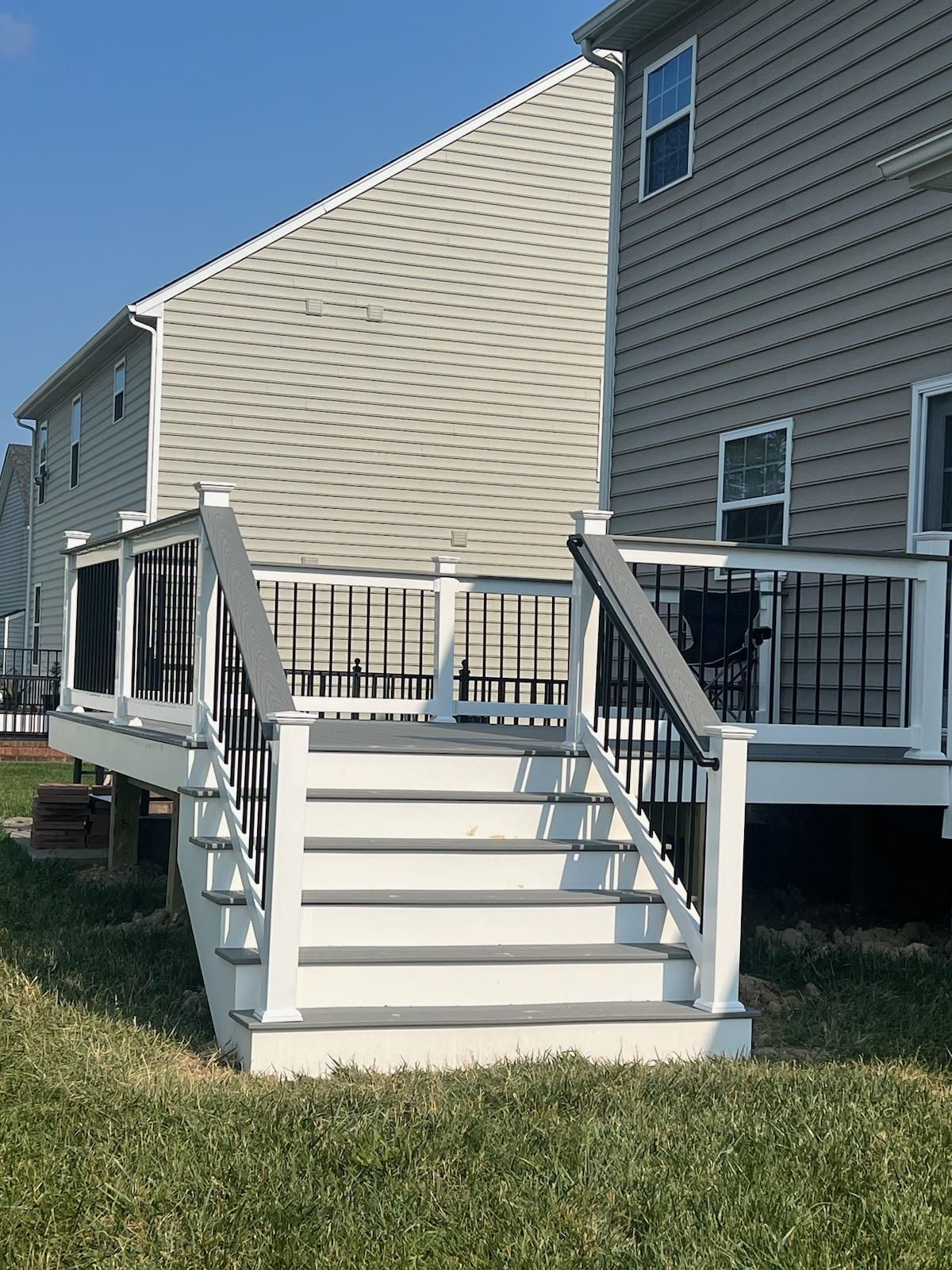 Gray and white deck with stairs leading to a house with gray siding, set in a grassy yard under a clear sky.