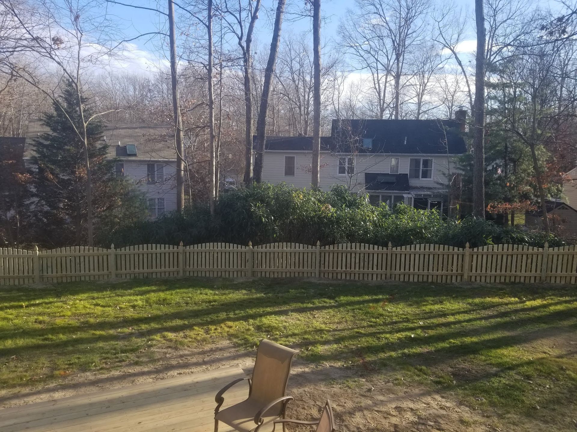 Lawn with a light brown picket fence in front of a house, and tall bare trees in the background.