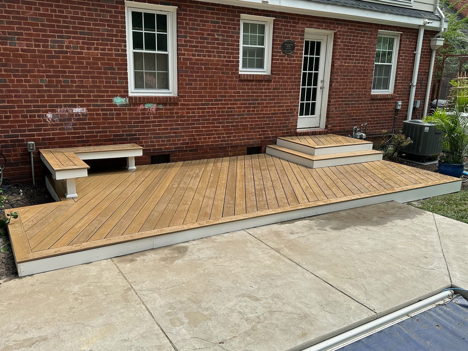 A wooden deck with bench and steps against a brick building, over a concrete patio.