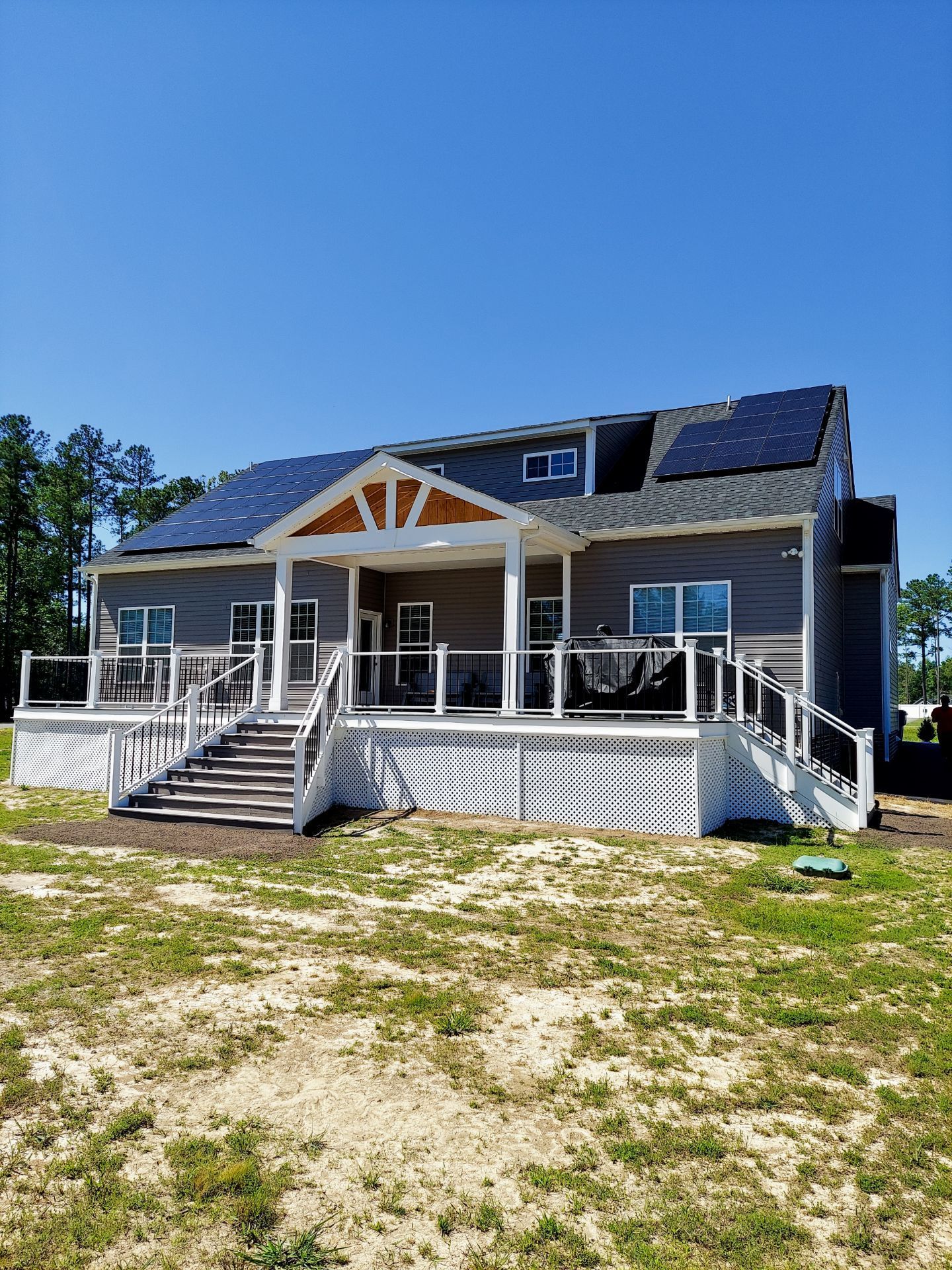 Large two-story house with gray siding, a deck, and solar panels on the roof against a blue sky.