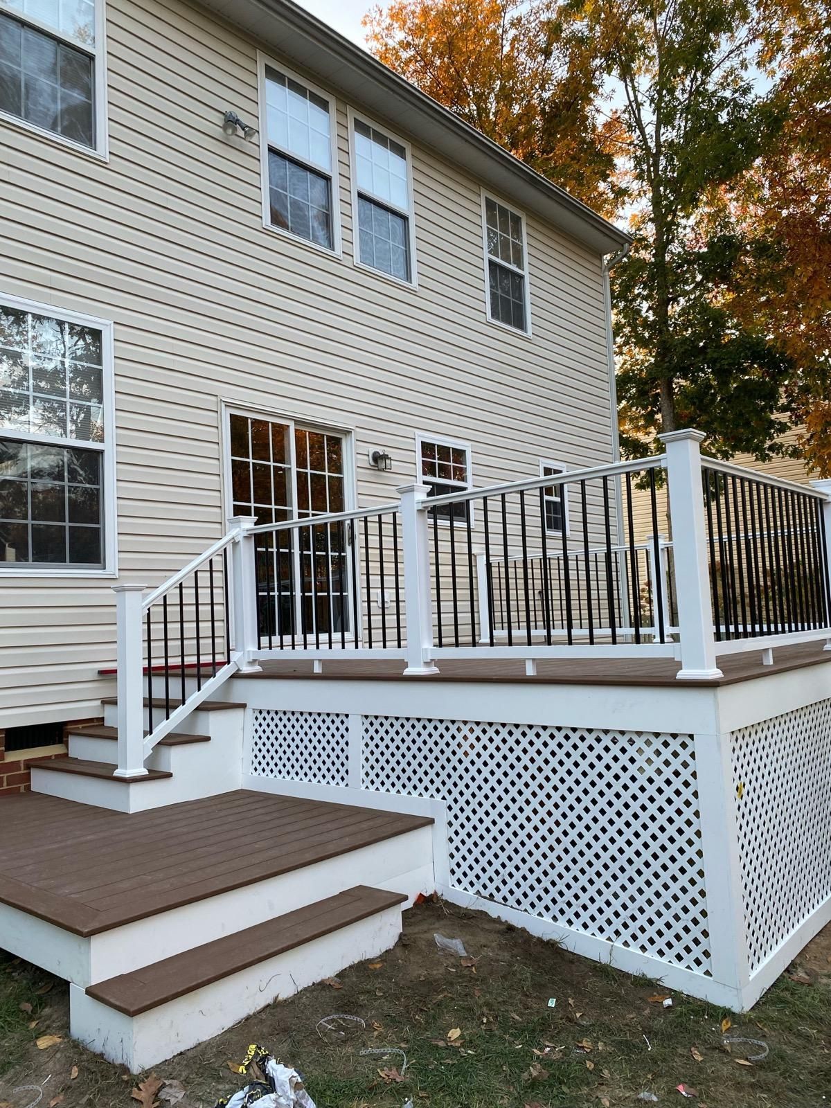 Back of a two-story beige house with a brown deck, white railing, and lattice skirting; steps lead down to the yard.