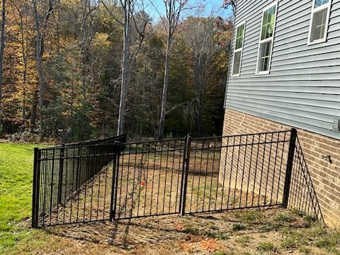 Black metal fence enclosing a small yard beside a house, with a wooded area in the background.
