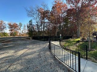 Gravel driveway and paved sidewalk with black fence leading to a treed area, against a bright blue sky.