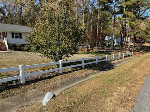 White fence along a grassy ditch, with a house and trees in the background.