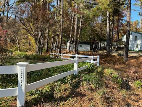 White fence in front of a yard with trees and buildings. Fall foliage with brown and green hues. Sunny day.