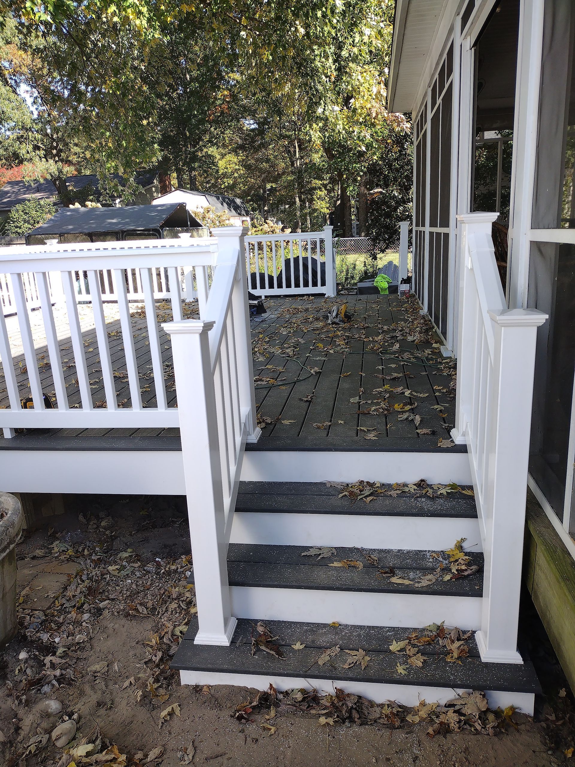 A white deck with black steps, covered in leaves, leads to a screened-in porch, surrounded by fall foliage.