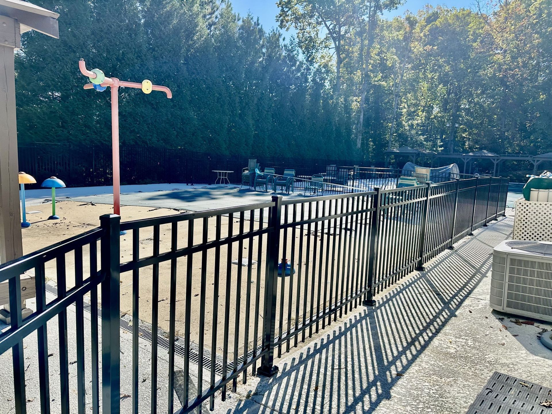 Black fence encloses a splash pad with water features, trees in background.