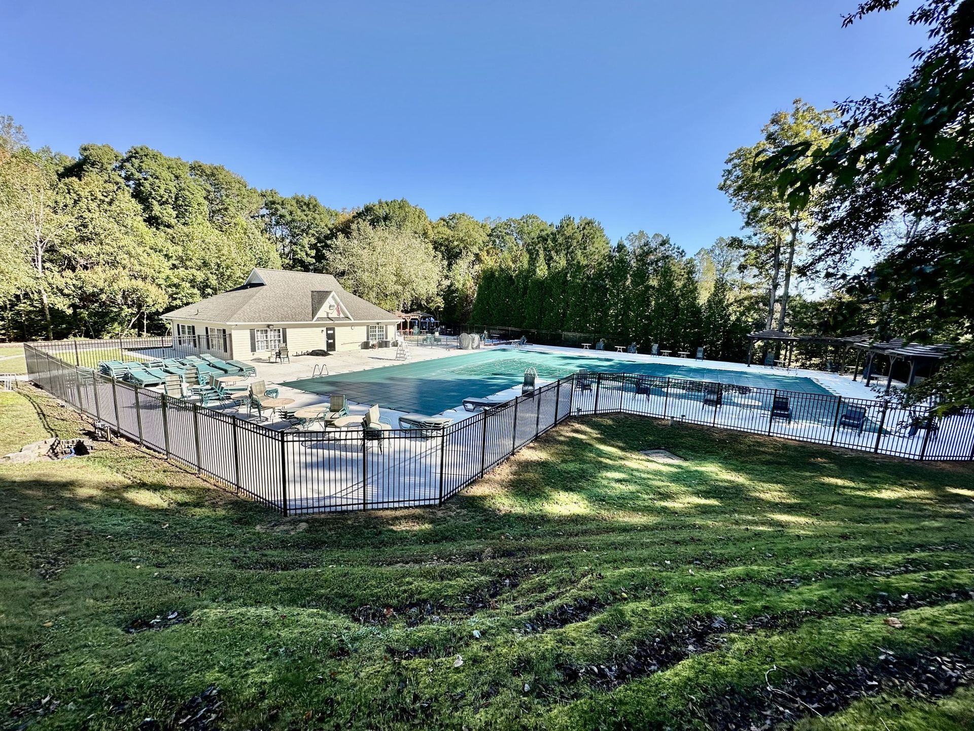 Pool covered with a green tarp, surrounded by fencing and trees, under a blue sky.