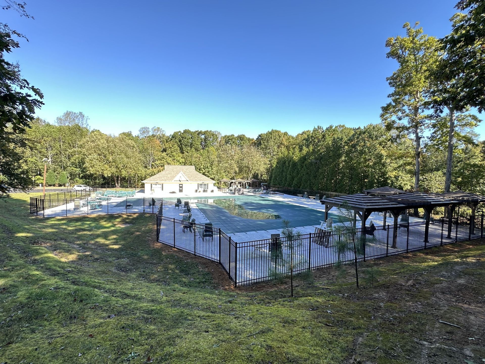 A fenced-in backyard pool with a gazebo, surrounded by trees and a white house.