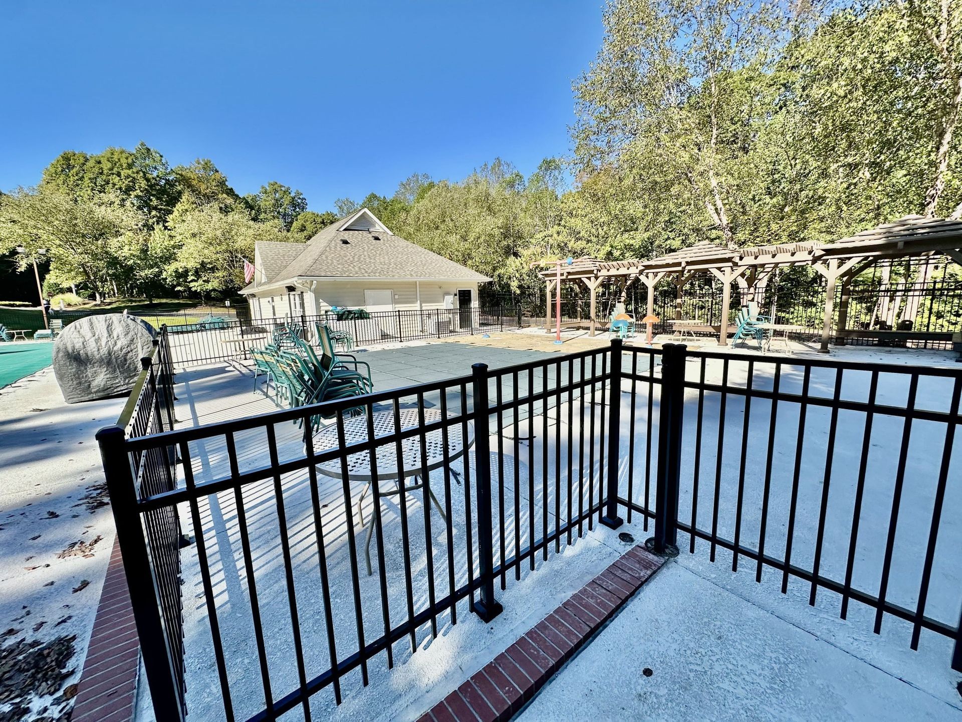 Black fence surrounds a pool area with a pool house, trees, and pergola under a blue sky.