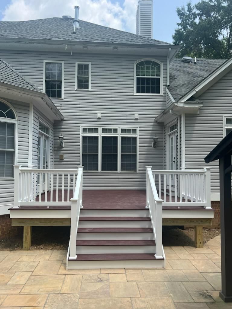 Back of a house with deck and stairs; gray siding, white trim, burgundy decking, and a stone patio.