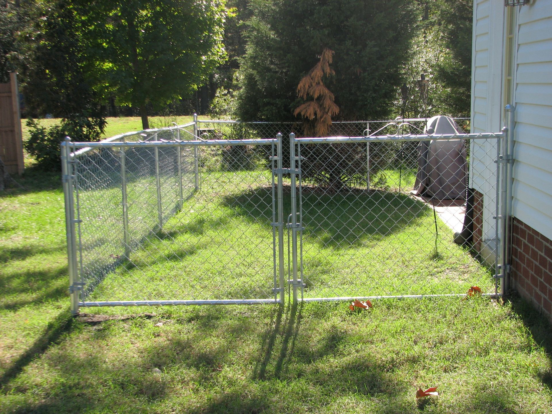 A chain-link fence encloses a small, grassy yard next to a brick-and-siding house. A gate is open.