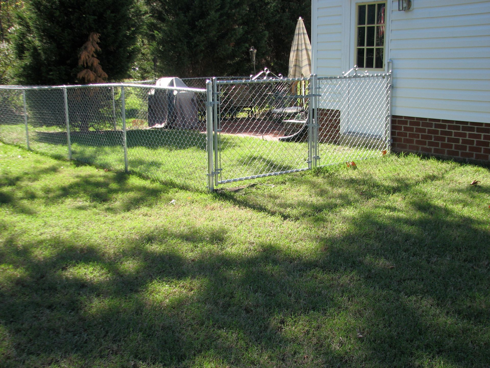Chain-link fence with gate in a yard. Sunny day, grass, house visible.