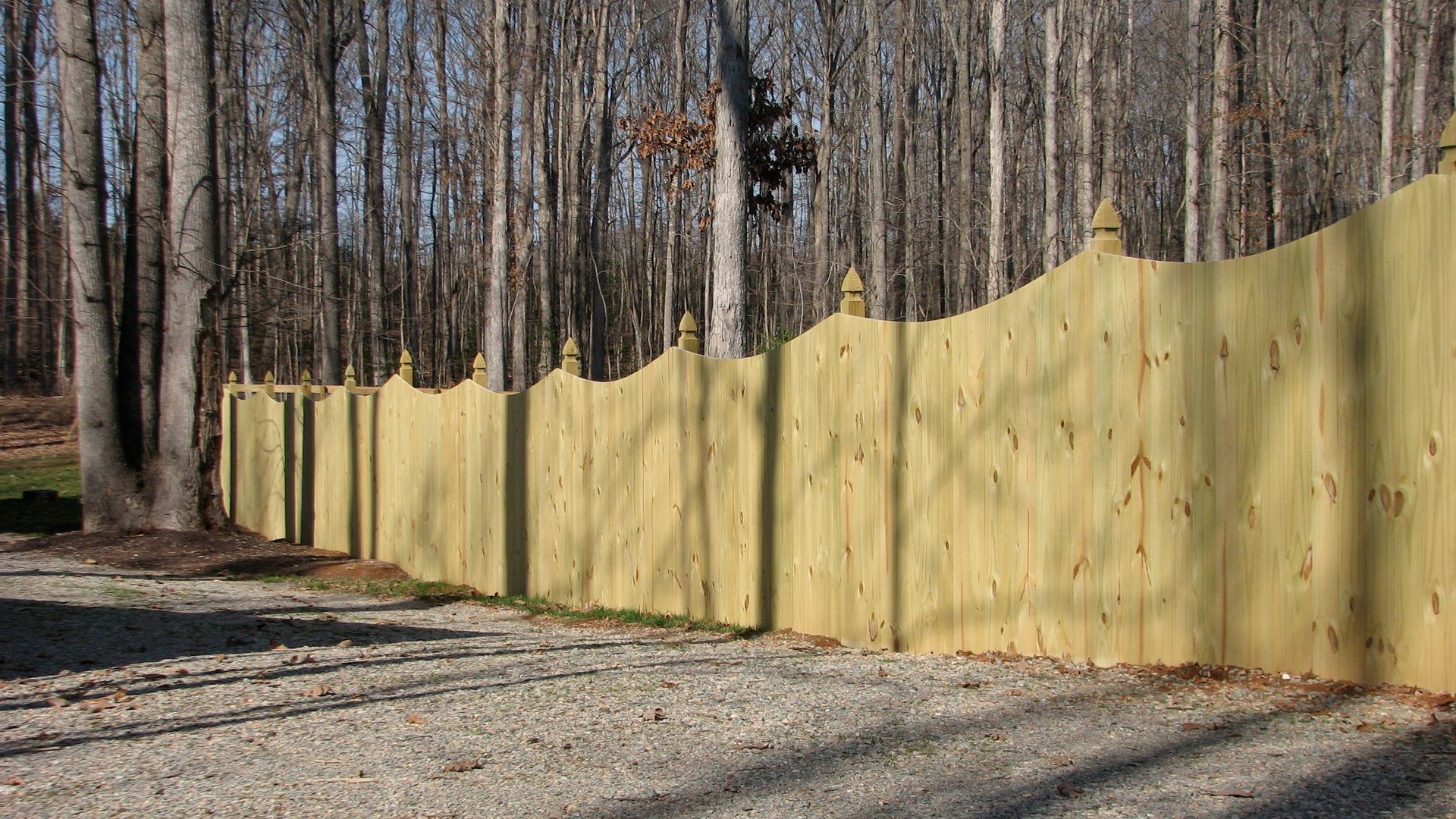 Wooden fence, painted yellow, with decorative tops, curves through a gravel area, trees in the background.
