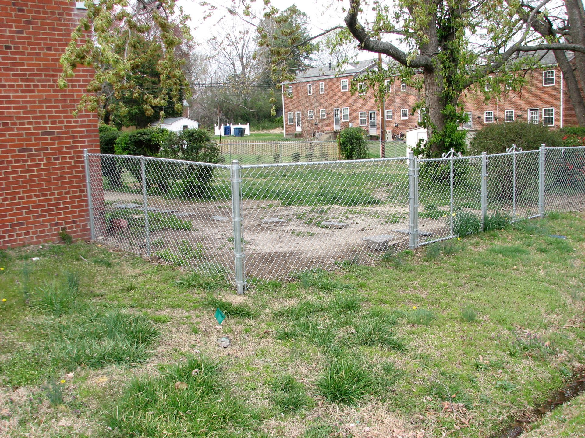 Chain-link fence encloses a bare dirt patch in a grassy yard, with brick buildings in the background.