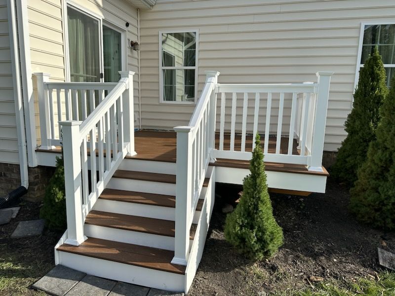 White railing and stairs lead to a deck on the side of a beige house, with small green bushes.