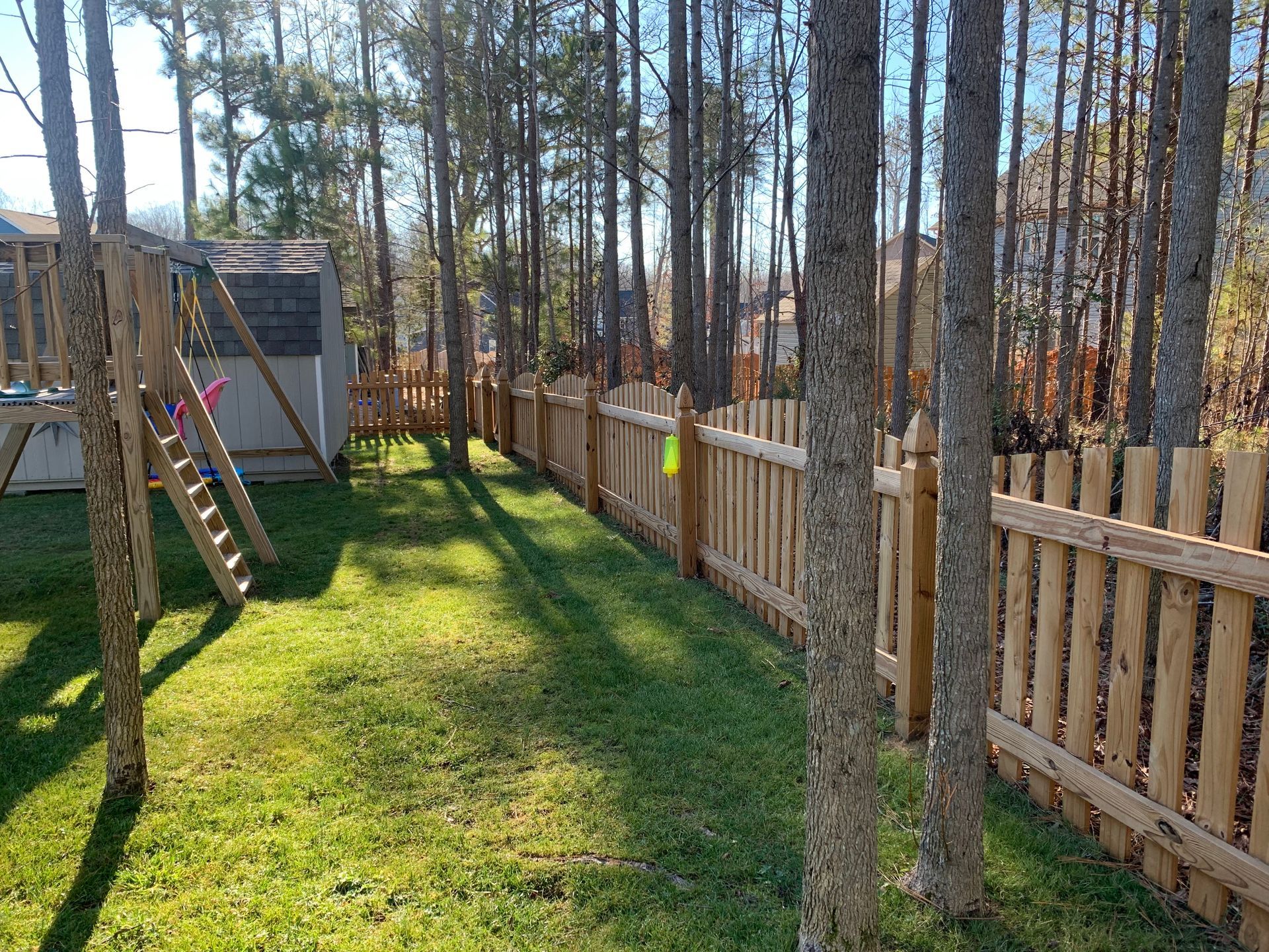 A sunny backyard with a wooden fence, green grass, trees, and a playset.