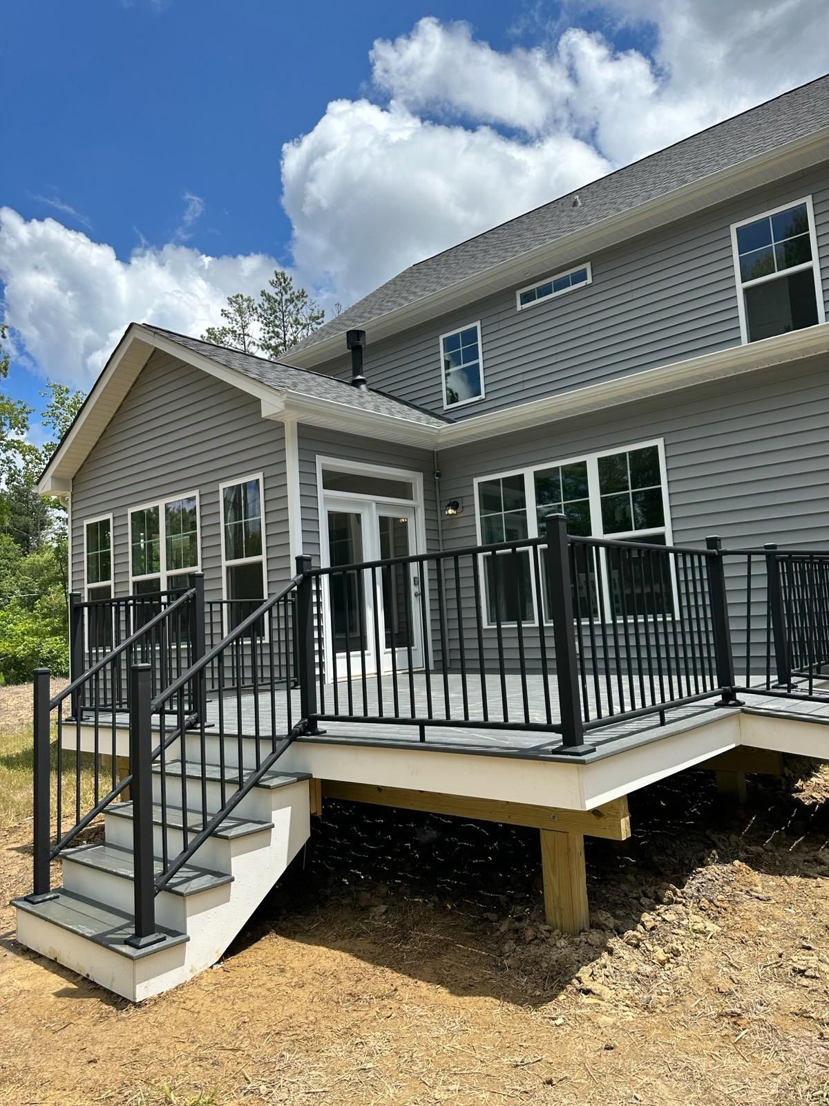 Gray house with black deck railing, stairs, and white trim. Blue sky and clouds.